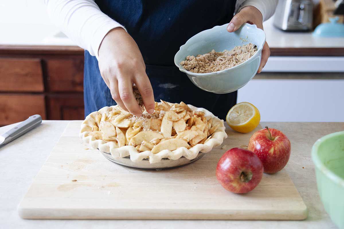 A woman puts crumble topping on a homemade Dutch apple pie just before baking.