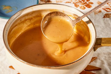 Angled view of a white pot of gravy sitting on a wooden trivet with a ladle with some gravy and all on a white and brown embroidered table cloth