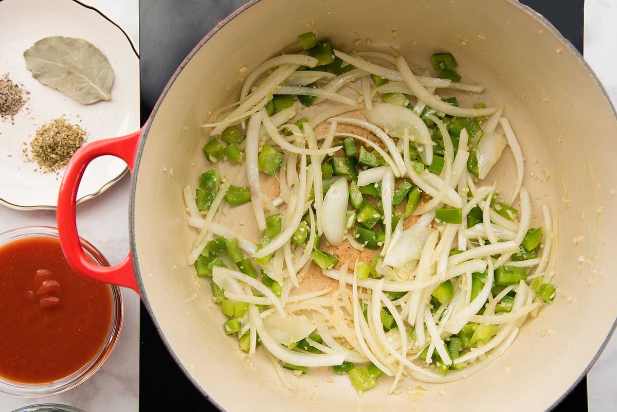 A dutch oven holds diced green peppers, sliced onion and garlic. On the counter next to the pot is a small glass bowl of tomato sauce and a scallopped plate with ground oregano, ground black pepper and a bay leaf.
