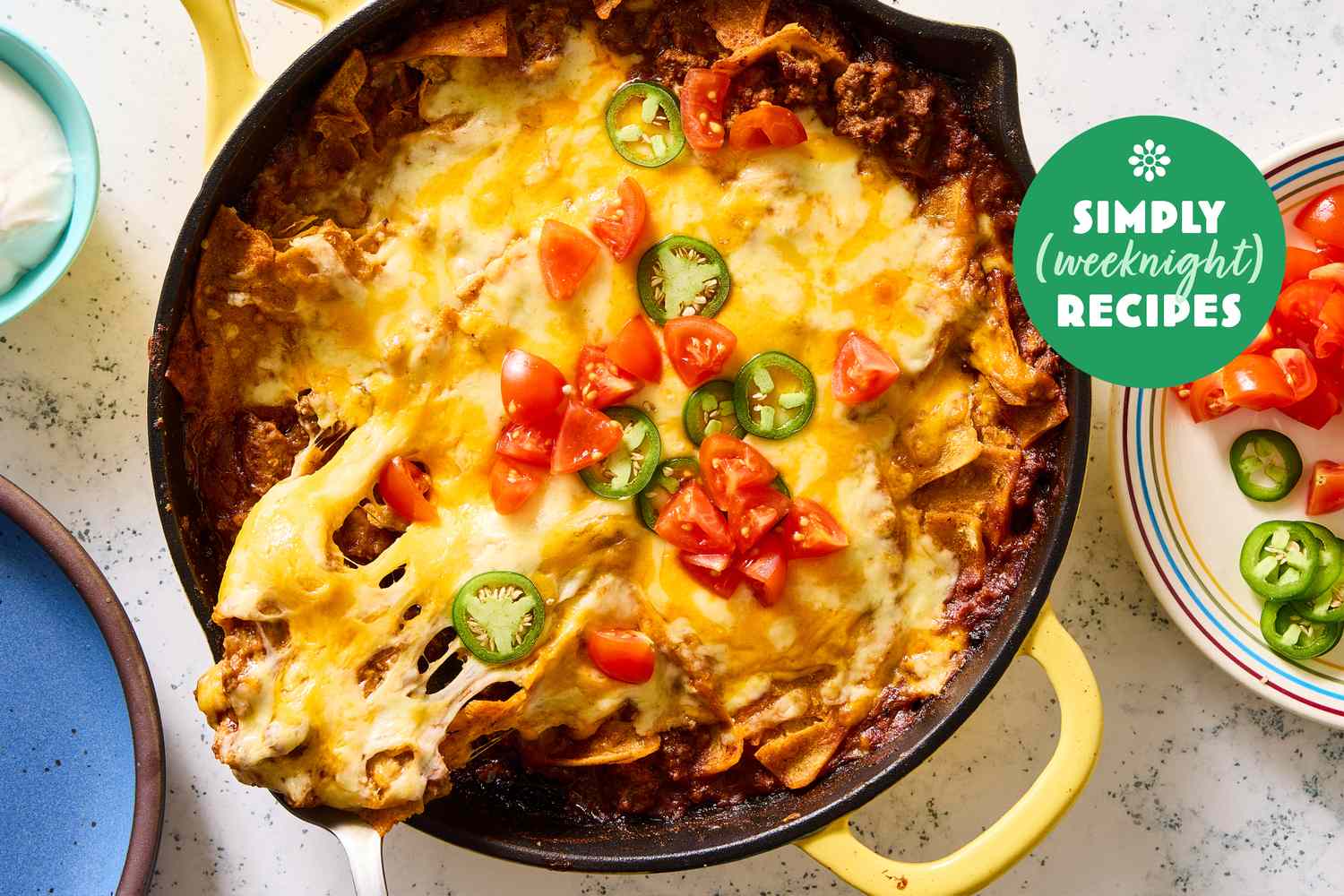 A skillet filled with cheesy nachos topped with diced tomatoes and jalapeos marked with a Simply Weeknight Recipes label