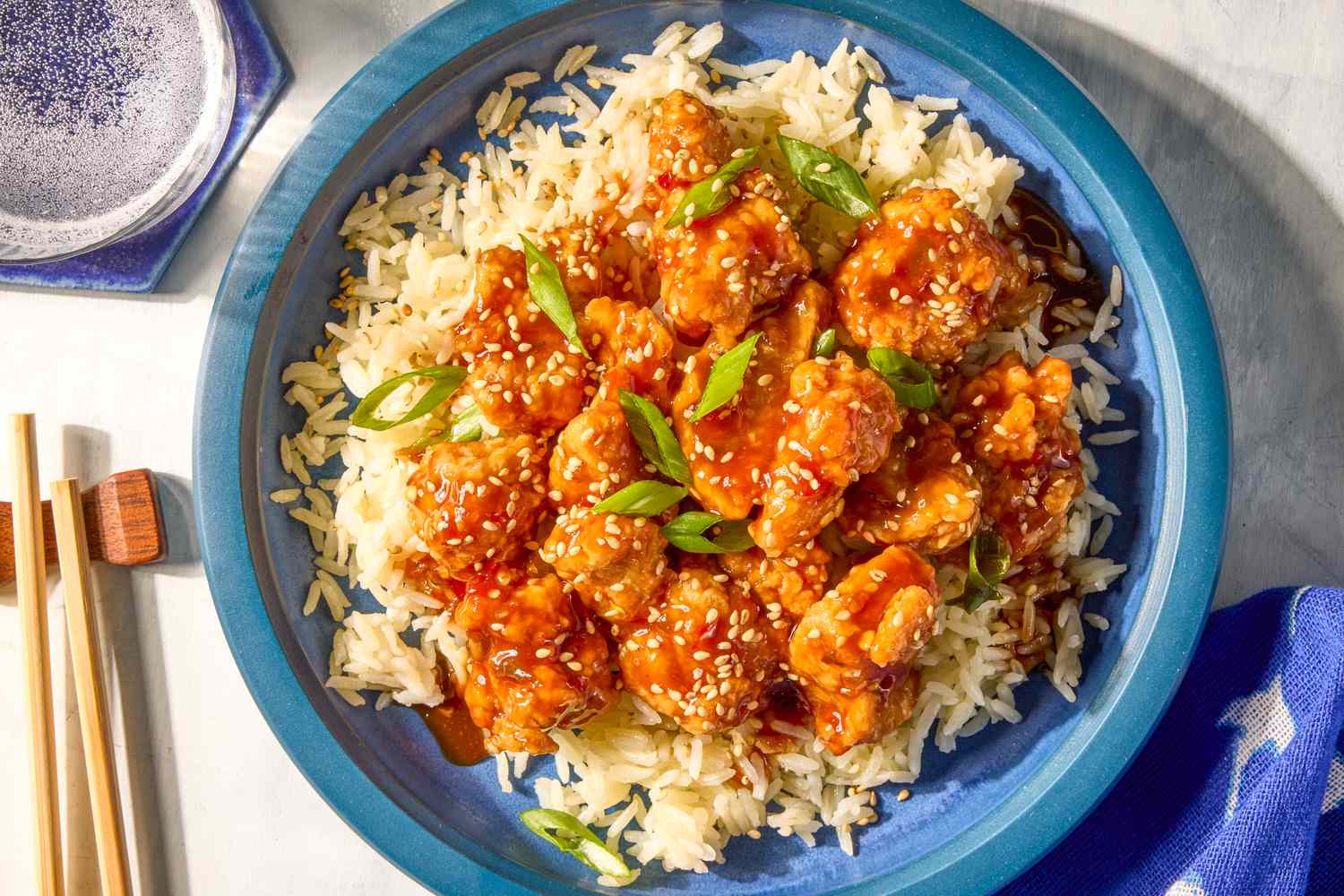 A plate of General Tsos chicken served on a bed of rice sprinkled with scallions and sesame seeds