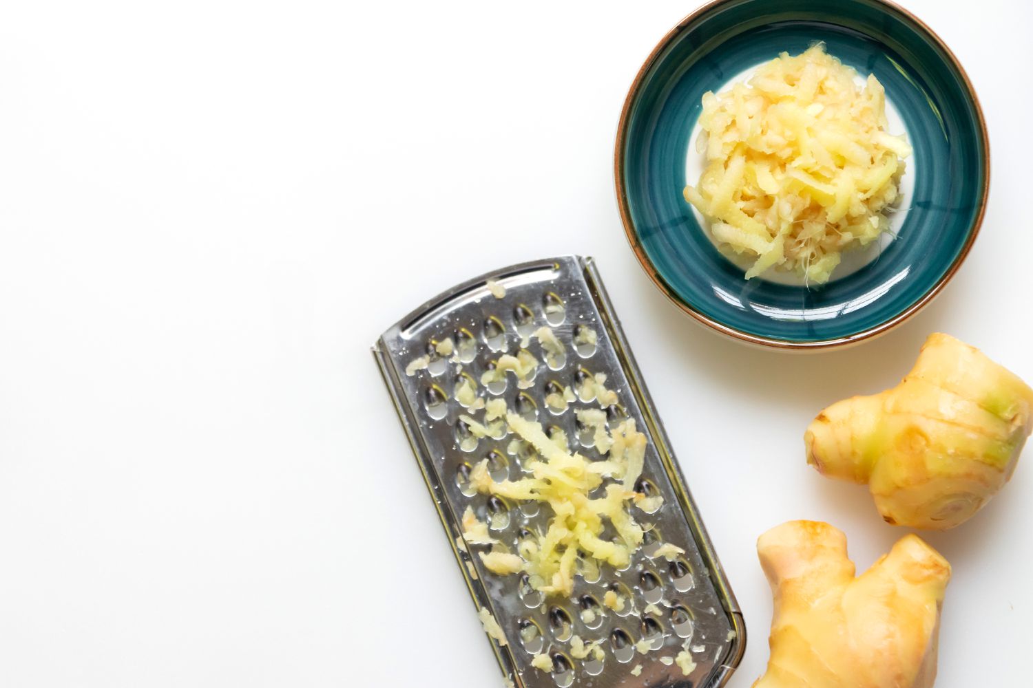 Peeled ginger, a handheld grater with grated ginger, and a small cup with grated ginger on a white backgroud