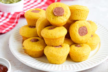Pile of Corn Dog Muffins Stacked on a Plate, and in the Background, a Red and White Checkered Kitchen Towel and Small Bowls of Toppings (Relish and Ketchup)