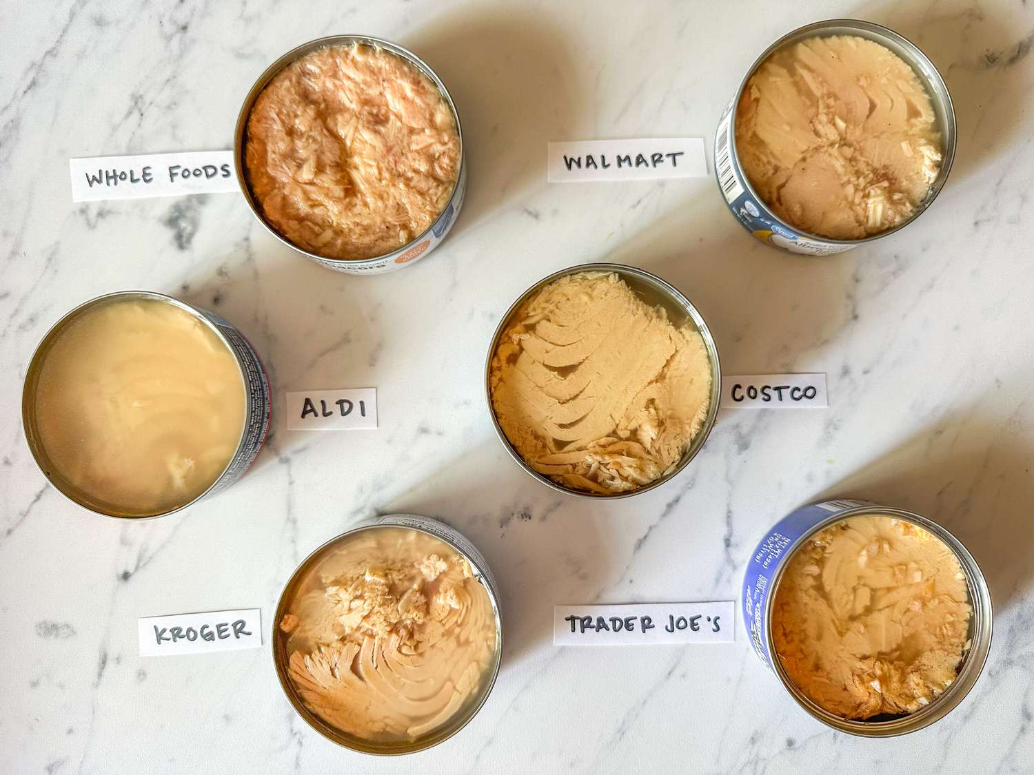Open cans of tuna from various brands labeled Whole Foods, Aldi, Kroger, Walmart, Costco, and Trader Joe's, displayed on a table