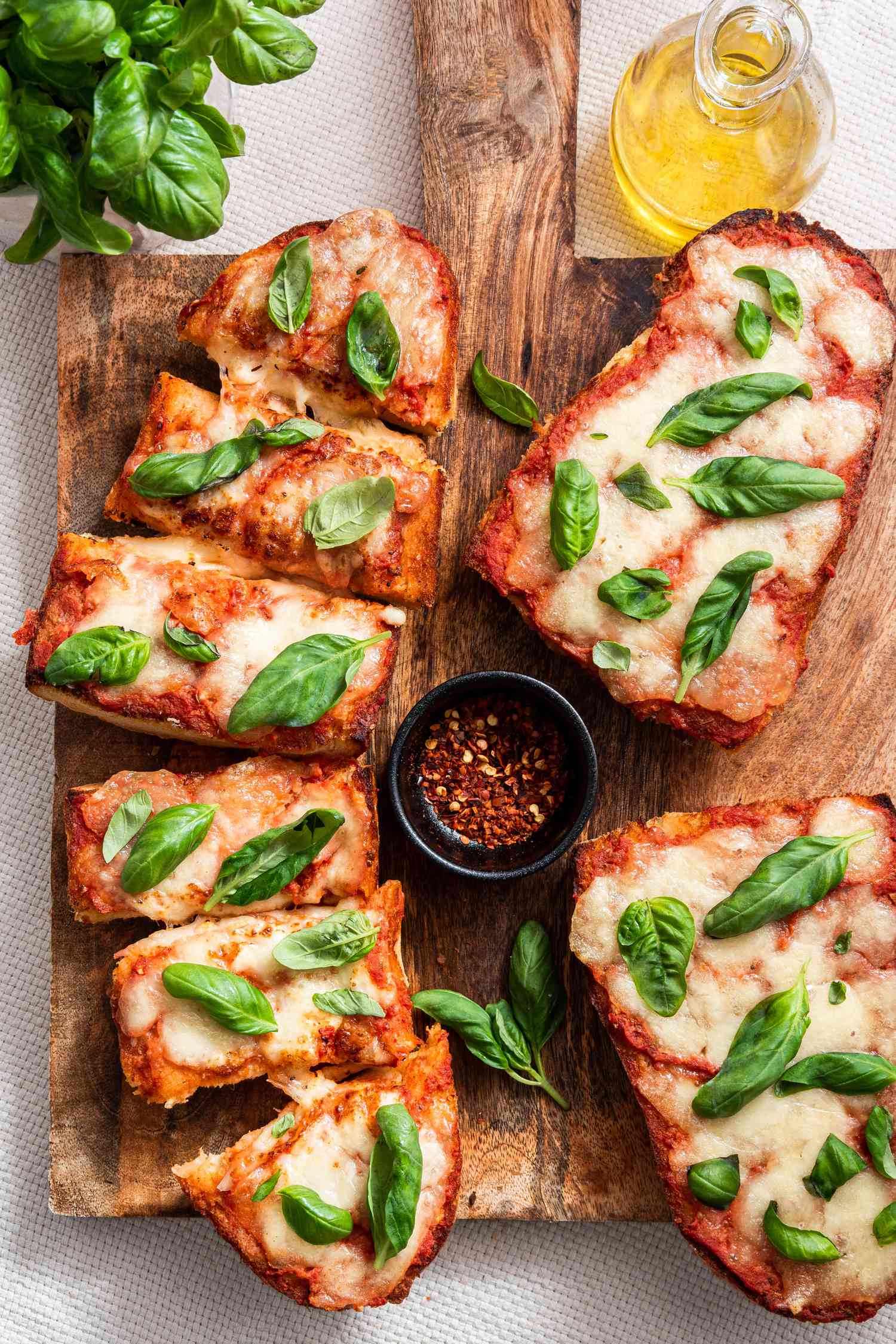 A Sliced French Bread Pizza Topped With Fresh Basil Next to More More Pizza (Unsliced) and a Small Bowl of Crushed Peppers on a Wooden Platter and on the Counter Next to It, an Olive Oil Dispenser and a Bushel of Fresh Basil 