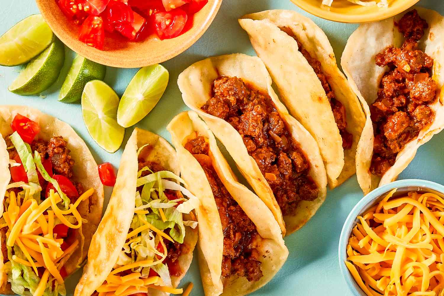 Crispy ground beef tacos on a plate a board with bowl of diced tomatoes, bowl of shredded cheddar cheese, and lime wedges