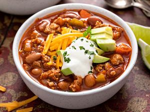 A bowl of chili topped with sour cream shredded cheese and avocado slices accompanied by lime wedges on the side