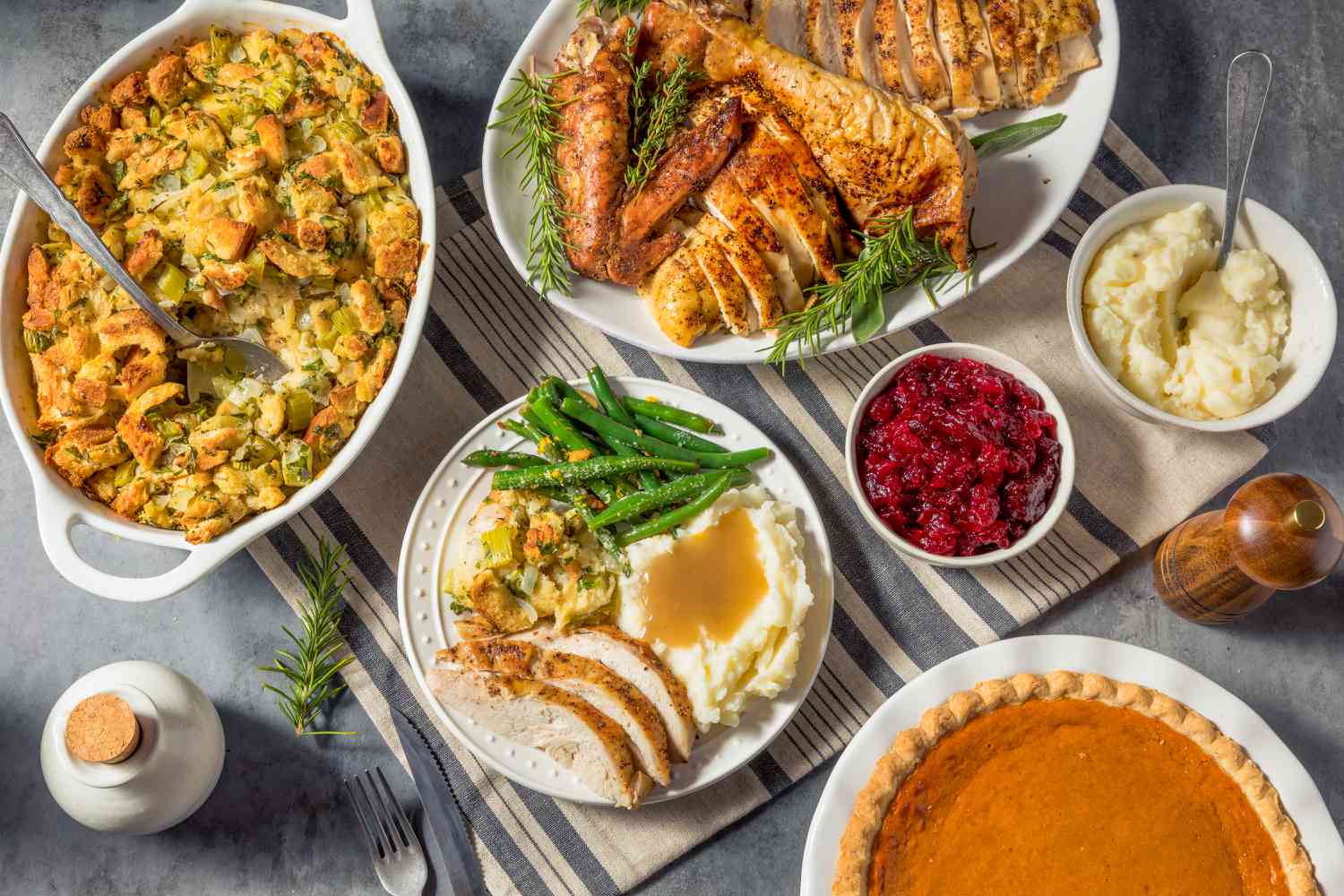 Overhead view of a spread of Thanksgiving foods including a plate of turkey, mashed potatoes, stuffing and green beans along with serving bowls for each, a bowl of cranberry sauce and a pumpkin pie