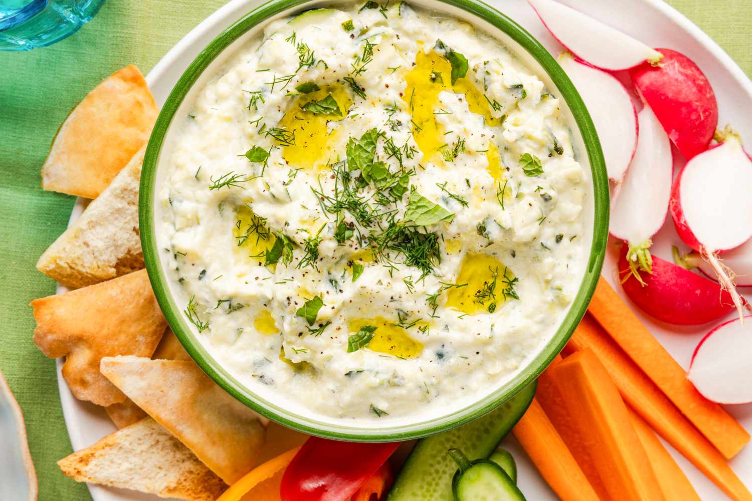 A serving bowl of creamy dip garnished with herbs and olive oil surrounded by fresh vegetables and pita bread for dipping
