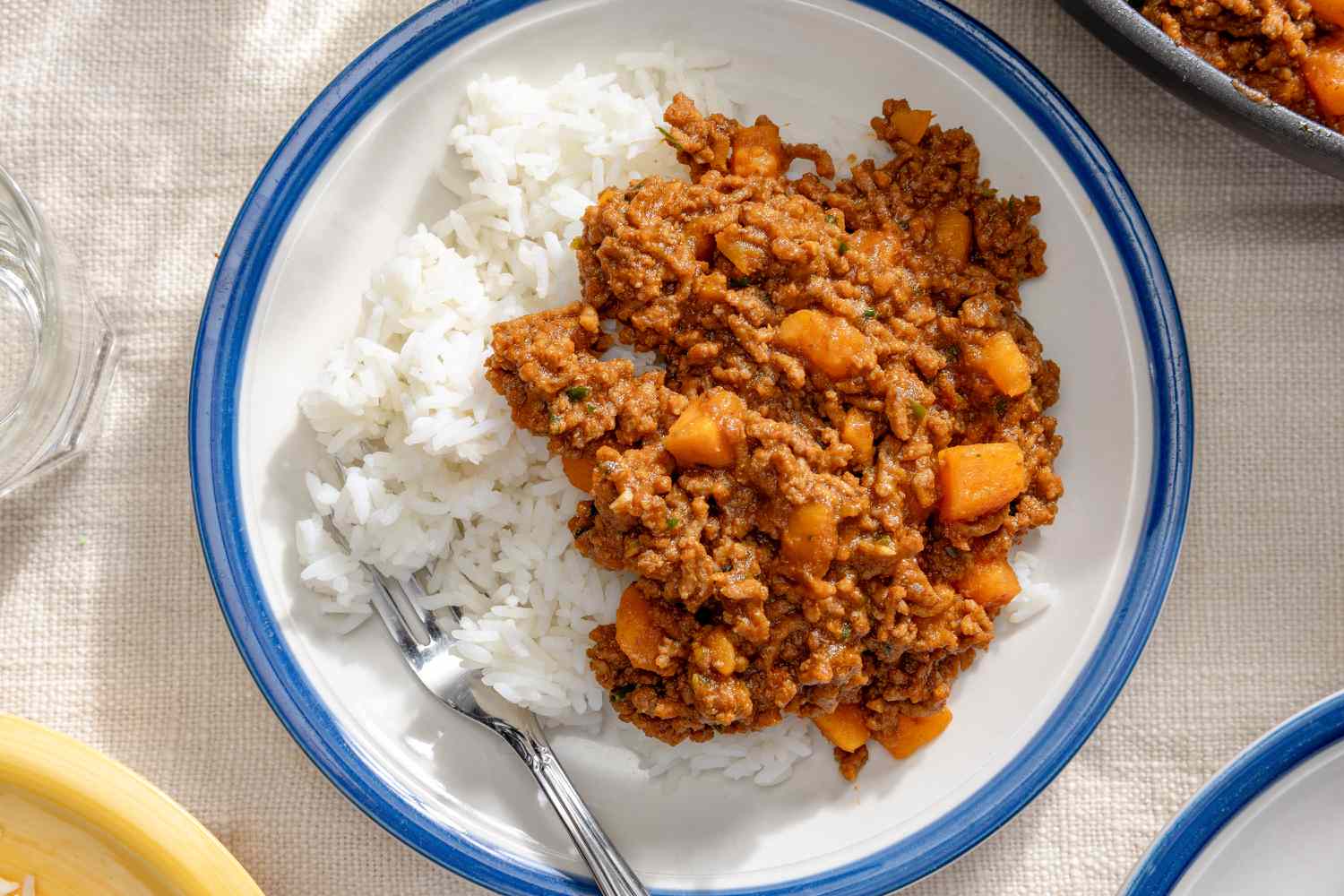 Overhead view of a white plate with a blue rim of picadillo over rice on a white table cloth