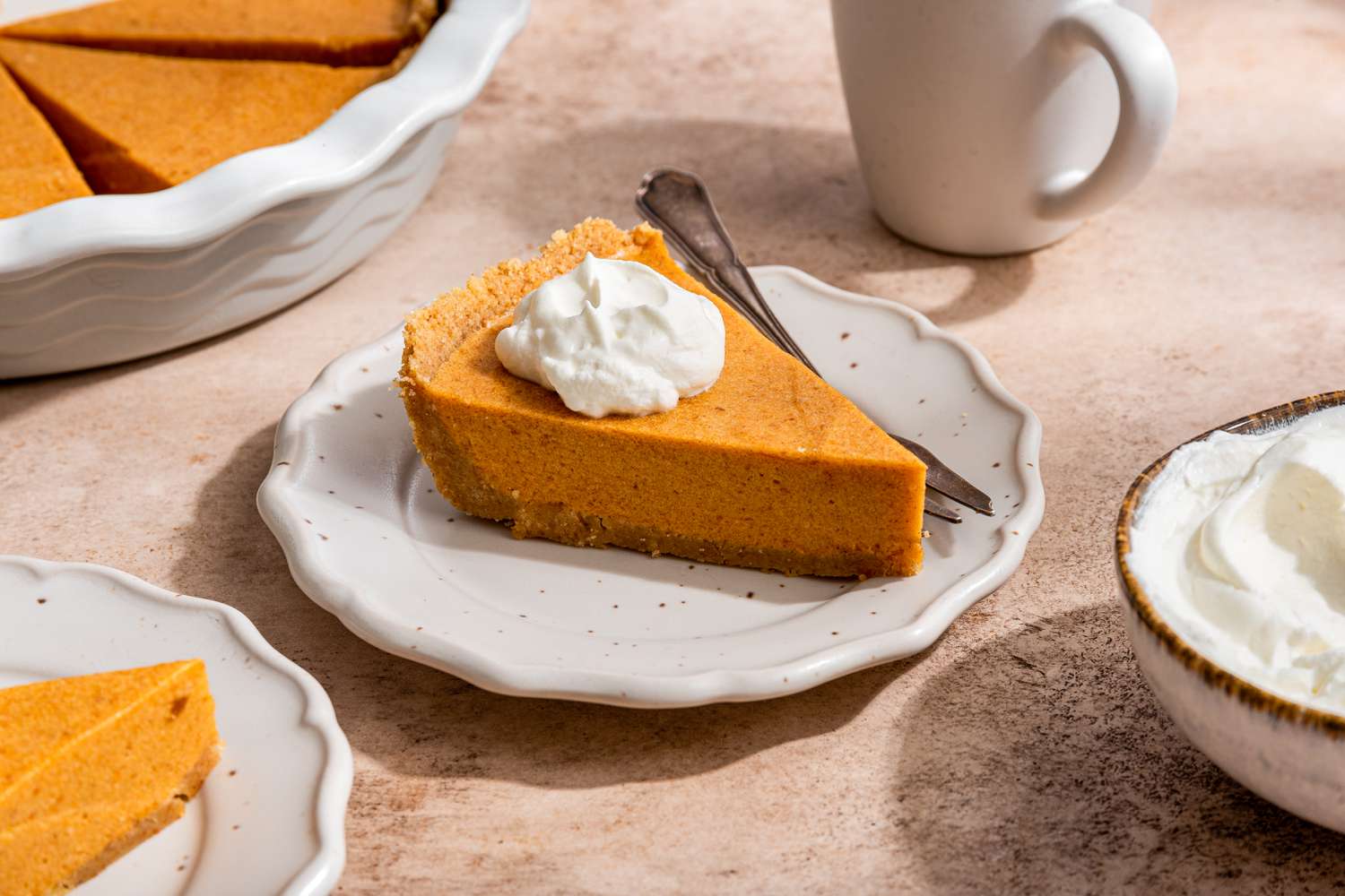 Slices of no-bake pumpkin pieon small plates topped with a dollop of whipped cream and a fork, a bowl of whipped cream, a cup of coffee, and a pumpkin pie in a pie pan