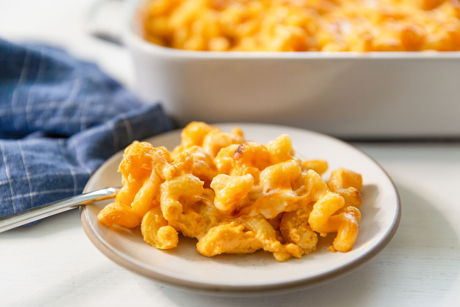 Side view of a small plate and a white baking dish of macaroni and cheese next to a blue table napkin