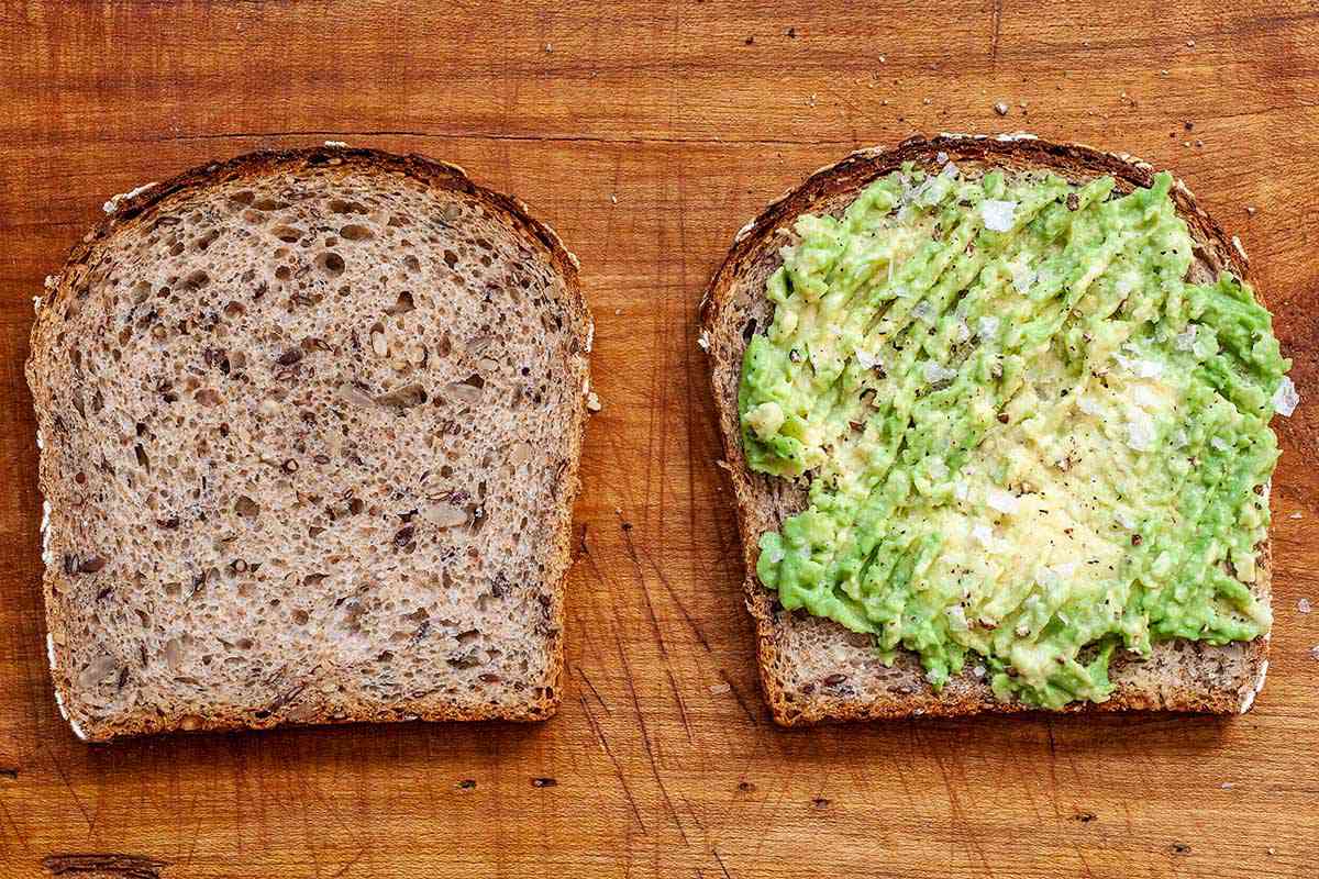 A slice of bread is topped with mashed avocado on a cutting board. A second slice is to the left.
