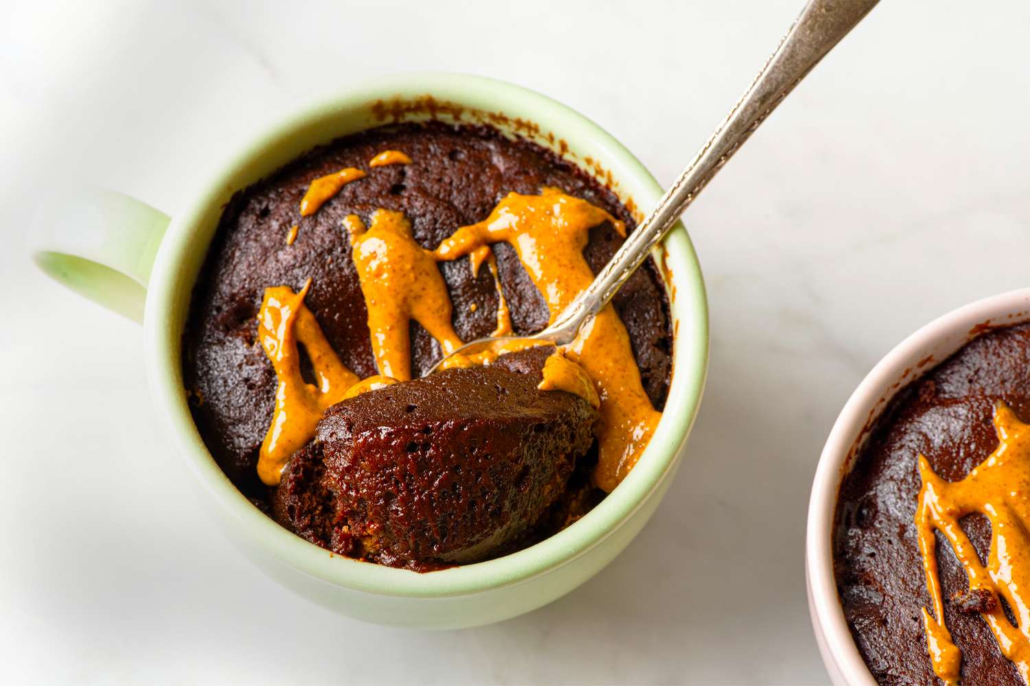 Chocolate mug cake topped with drizzled peanut butter a spoon scooping from one mug