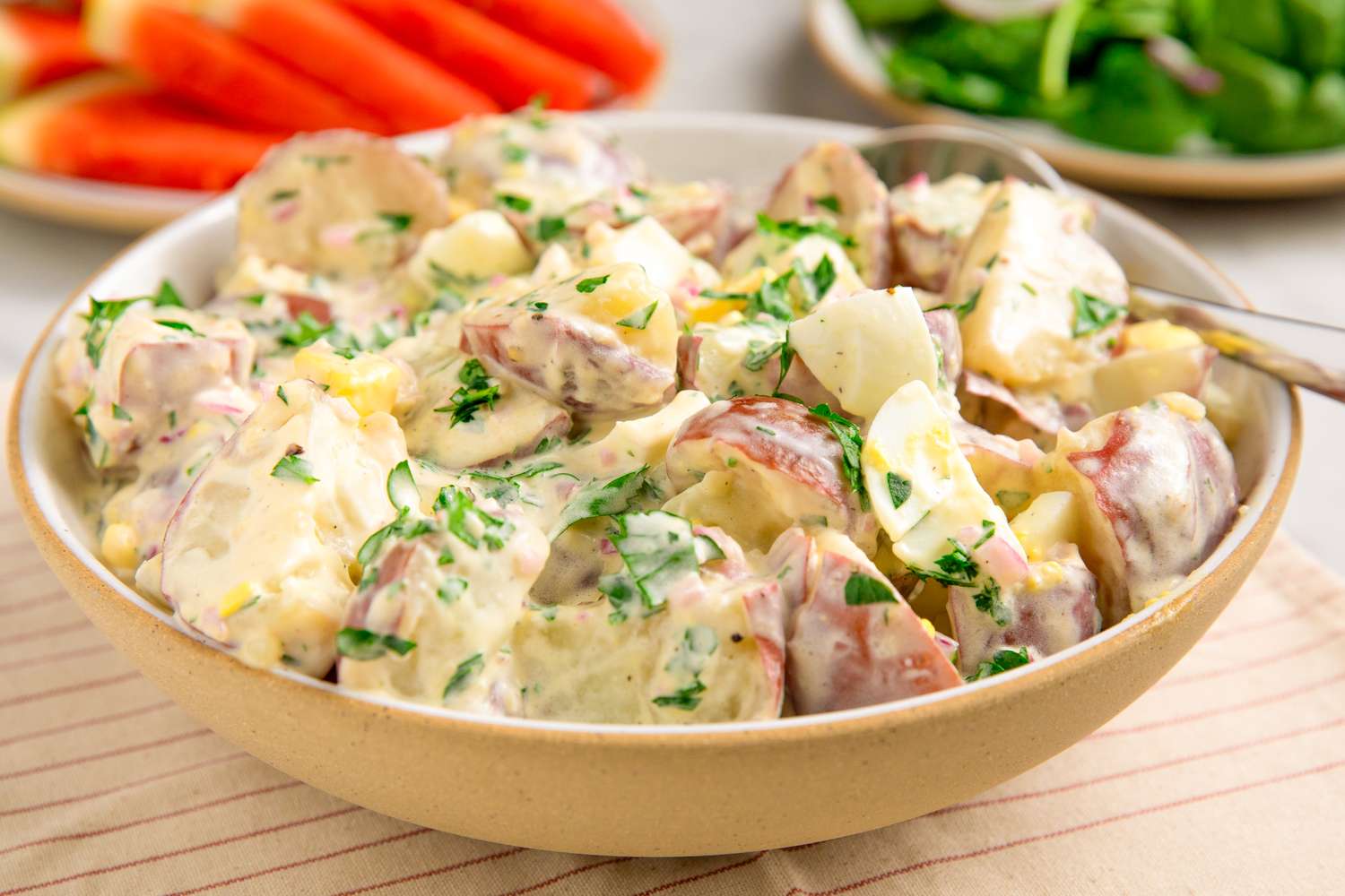A bowl of potato salad garnished with parsley sitting on a cloth with plates of vegetables in the background