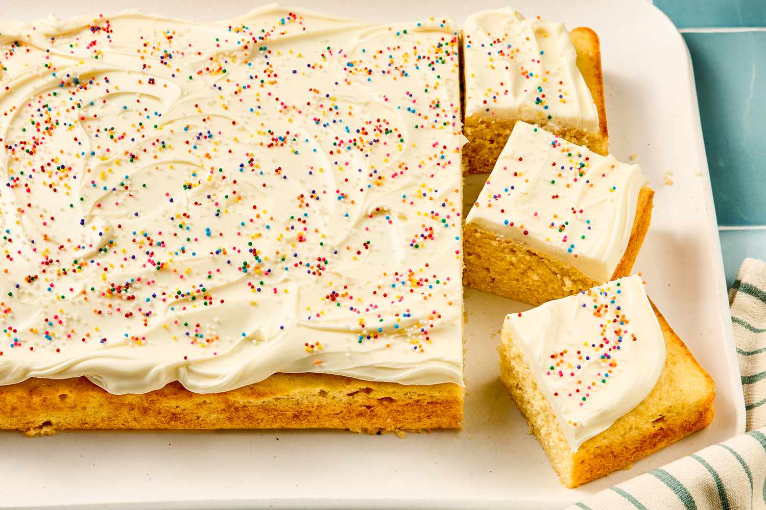 A rectangular vanilla cake with frosting and sprinkles, partially sliced into square pieces