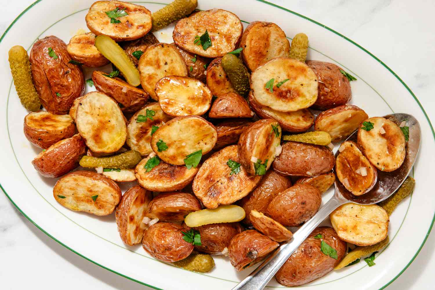 Overhead view of a serving spoon on an oval platter of roasted potato halves and pickles topped with parsley