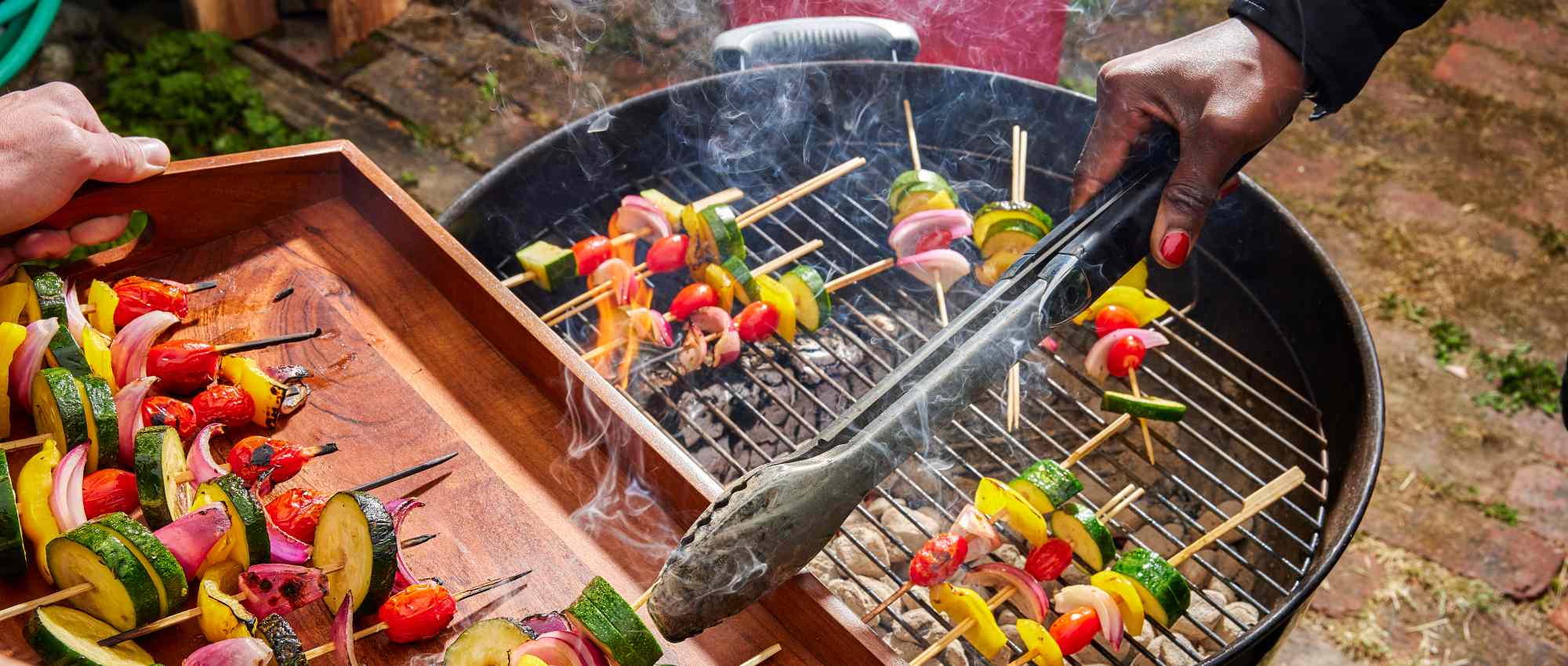 People grilling vegetable skewers on a barbecue grill, with a person holding tongs and another arranging skewers on a tray