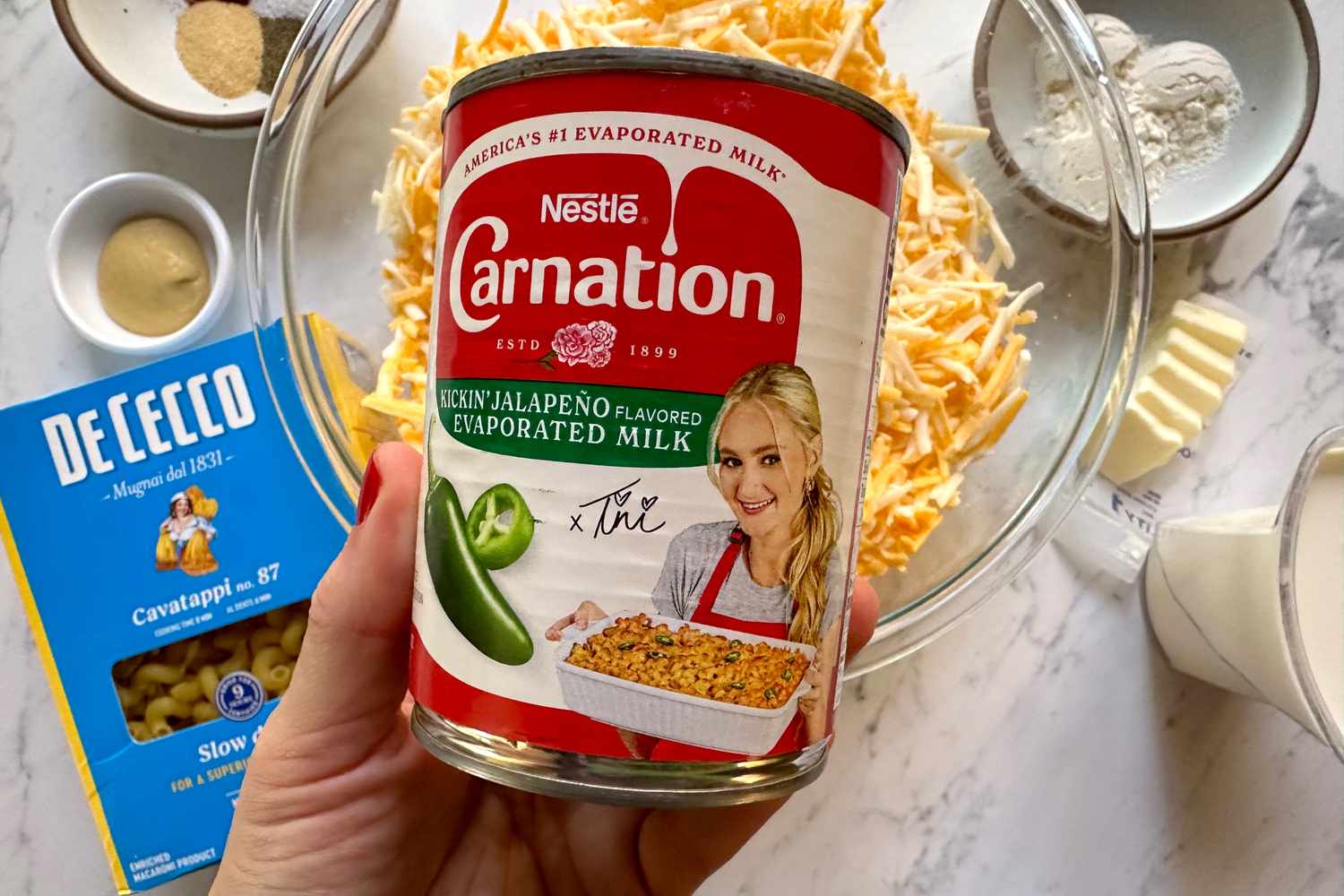 A hand holding a can of Carnation Kickin' Jalapeno flavored evaporated milk with other ingredients for mac and cheese on a table