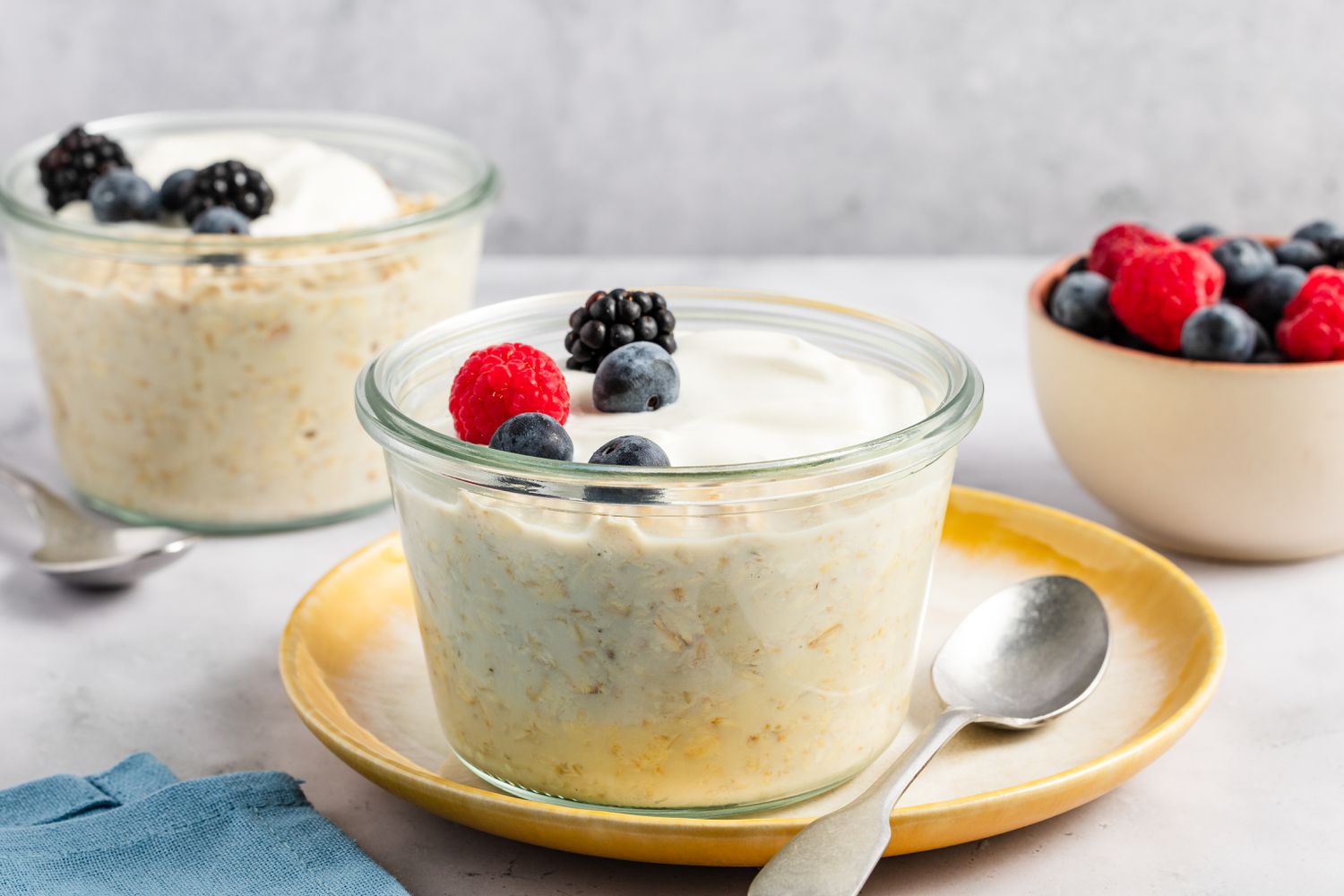 Two glass ramekins with overnight oats, topped with fresh berries, with a small bowl of berries in the background