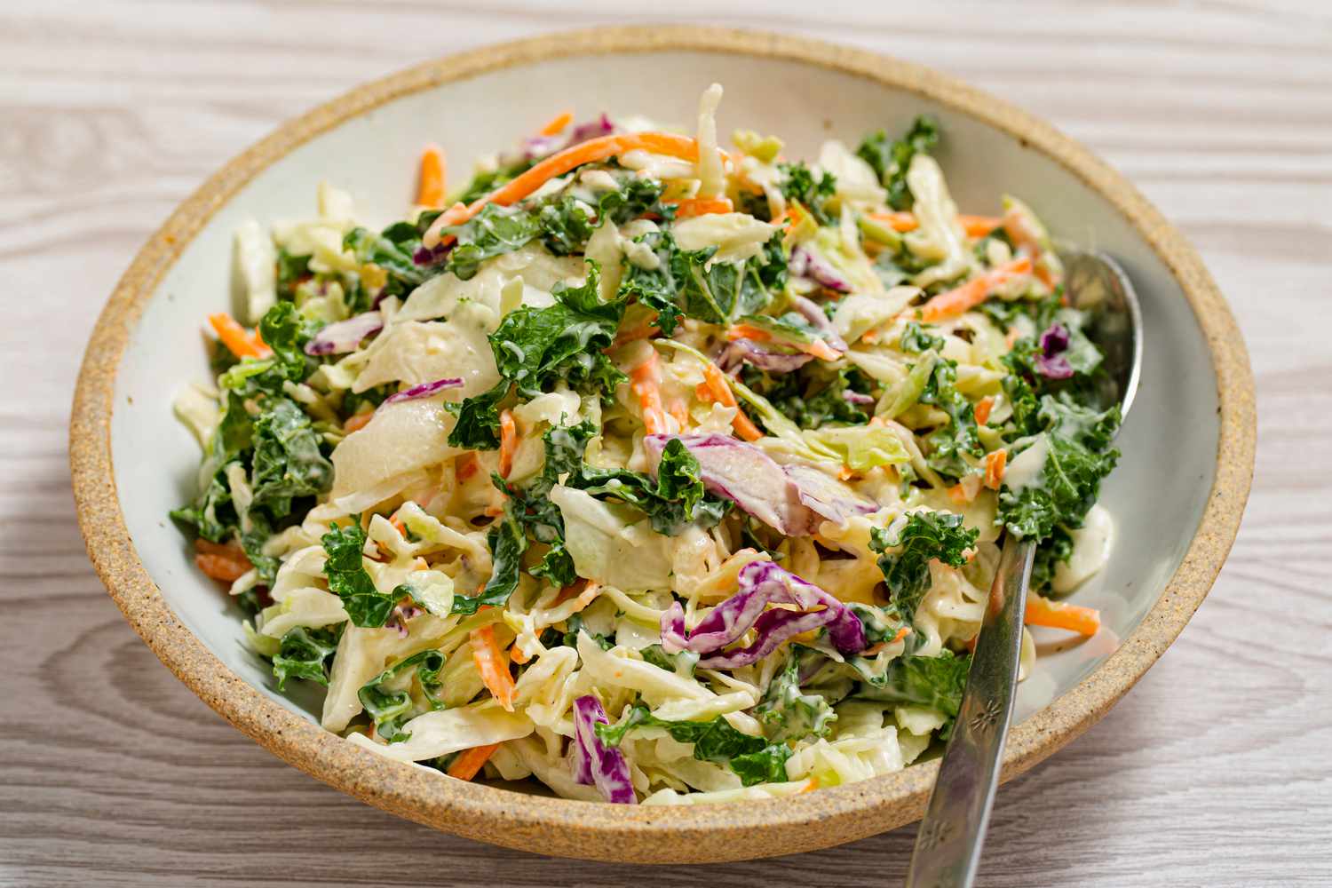 A bowl of coleslaw with various vegetables, placed on a wooden surface with a spoon