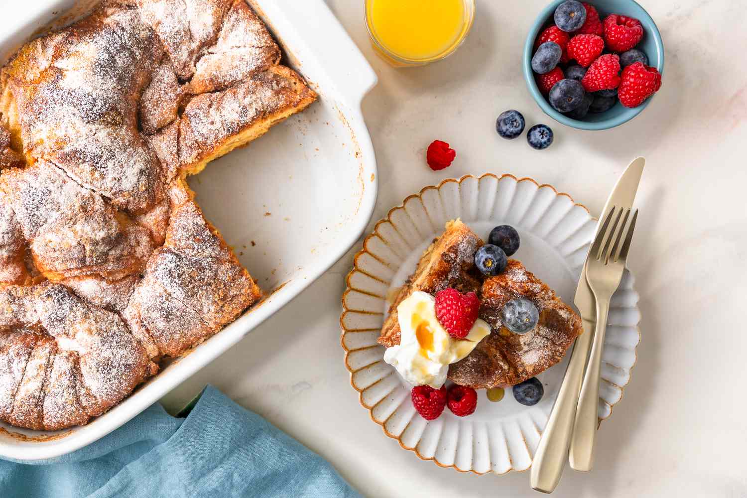 Overhead view of a white baking dish of Croissant French Toast casserole with a slice removed and placed on a small plate with a knife and fork along with a glass of orange juice and bowl of fruit