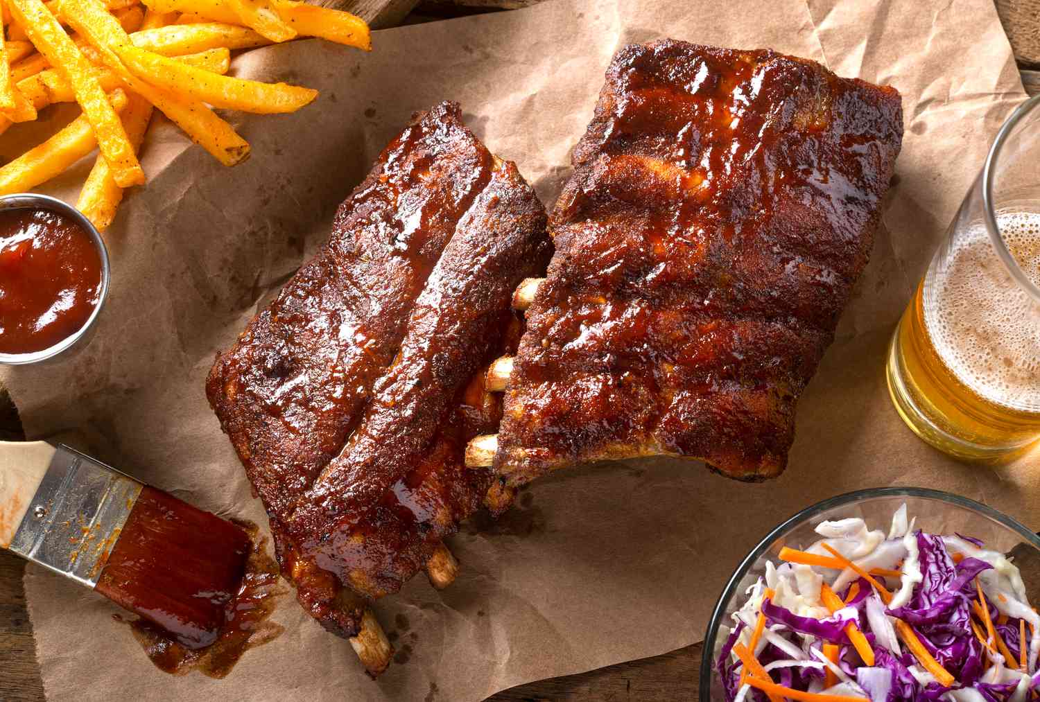 rack of glazed ribs on parchment paper at a table setting with a basket of fries, bowl of coleslaw, and a glass of beer 