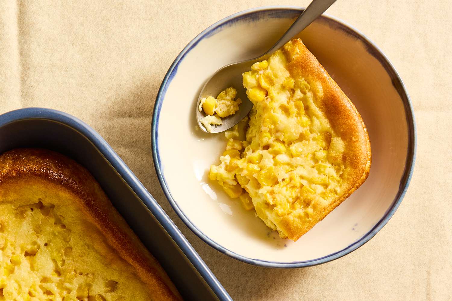 Overhead view of a white bowl with a serving of corn soufflé and a spoon next to a blue baking dish if An image of a dark blue baking dish of soufflé