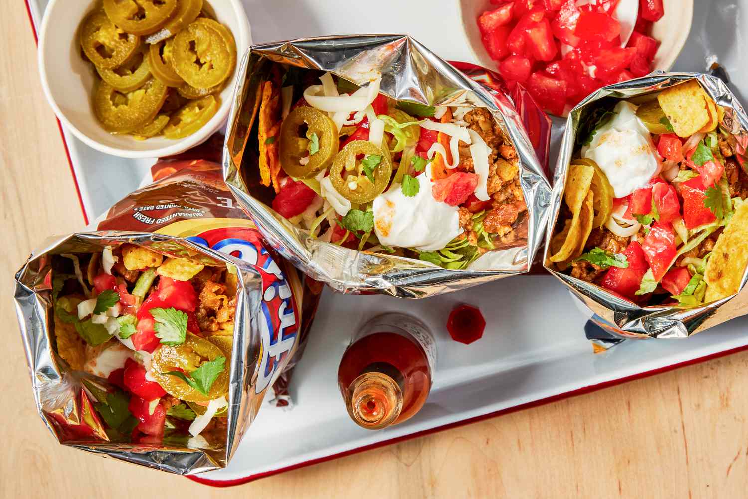 Multiple bags of walking tacos on a tray with a bowl of diced tomatoes, bowl of jalapenos, and a bottle of hot sauce