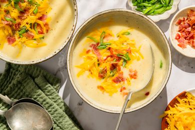 Overhead view of two bowls of potato soup topped with bacon, shredded cheese and green onions next to a bowls of bacon, green onions and shredded cheese