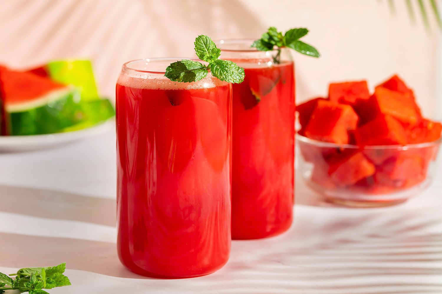 Two glasses of watermelon smoothie garnished with mint leaves slices of watermelon and diced watermelon in the background