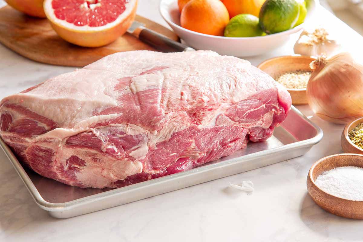 A baking tray with an uncooked pork roast inside. Wooden bowls of spices, as well as an onion, garlic and citrus fruit are placed to the right and above the pork.