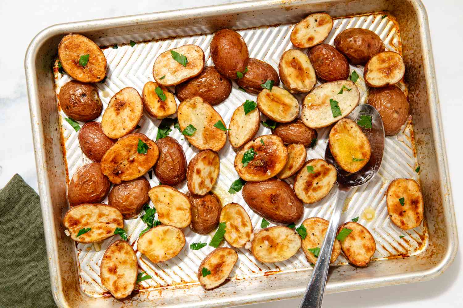 Overhead view of a serving spoon on a metal sheet pan of roasted potato halves topped with parsley