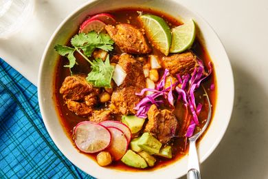 A bowl of pozole rojo garnished with lime wedges radish slices cabbage cilantro and pieces of pork placed on a blue tablecloth