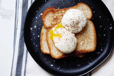 Two poached eggs on top of two pieces of stacked toast. One egg has a slightly runny yolk. Salt and pepper are sprinkled on the eggs, toast and black plate.