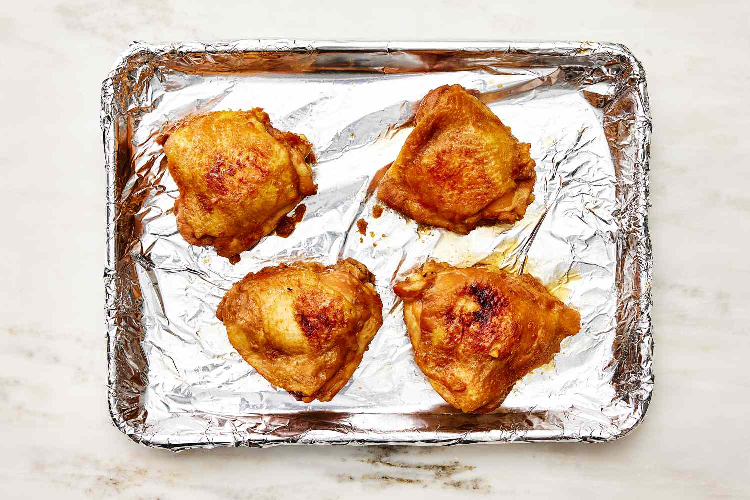 Overhead view of a foil lined baking sheet of four soy chicken thighs on a stone countertop for Shoyu Chicken recipe