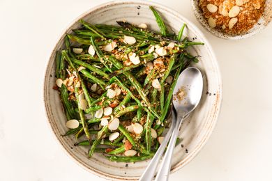 Bowl of broiled green beans topped with almond slices and crumbs, two spoons placed inside the bowl