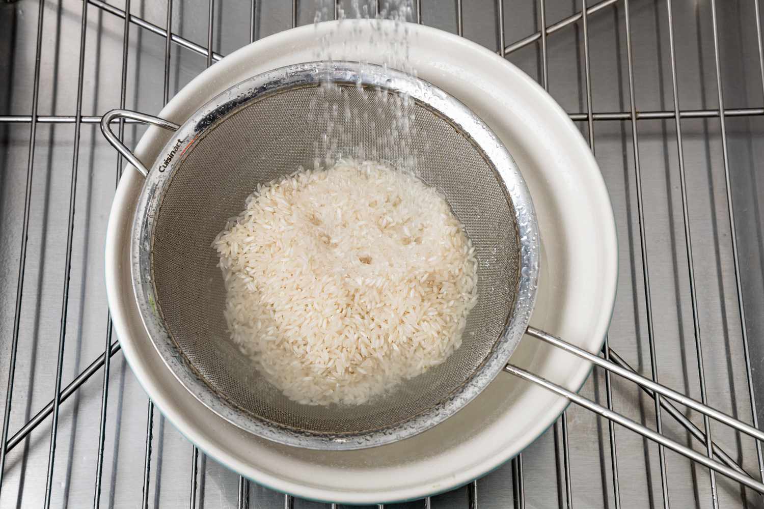 In the Kitchen Sink, White Rice in a Colander over a Bowl with Water from the Faucet Running for Microwave Rice Recipe