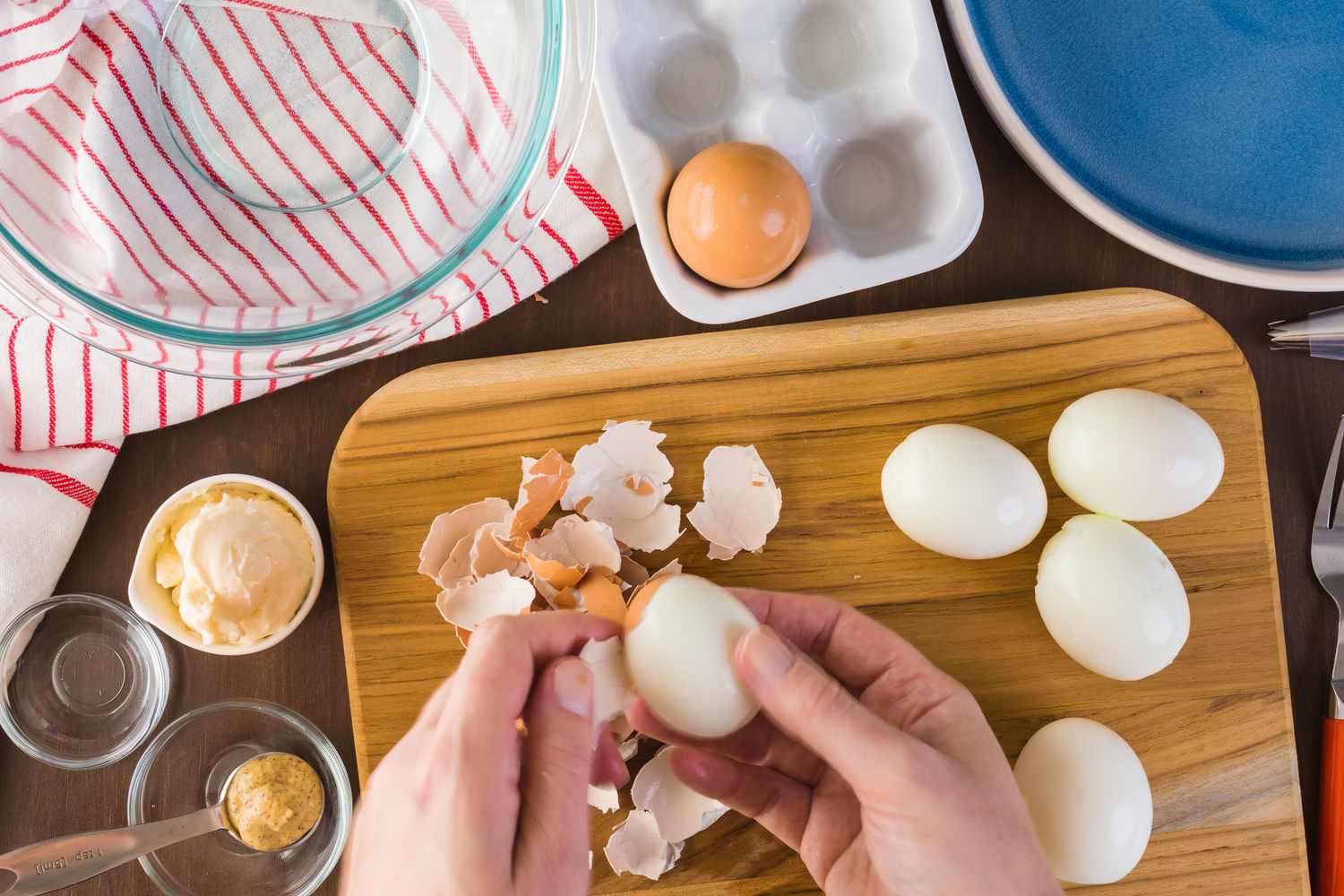 Overhead view of hands peeling boiled eggs on a wooden cutting board next to various bowls and plates and ingredients