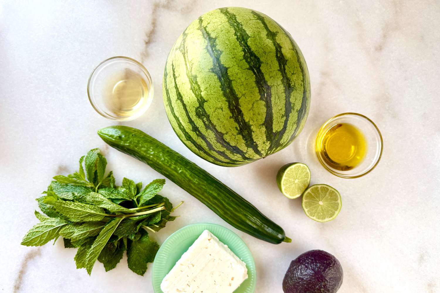 Ingredients arranged for a watermelon salad, including a watermelon, cucumber, mint, lime, feta cheese, oil, and avocado on a countertop