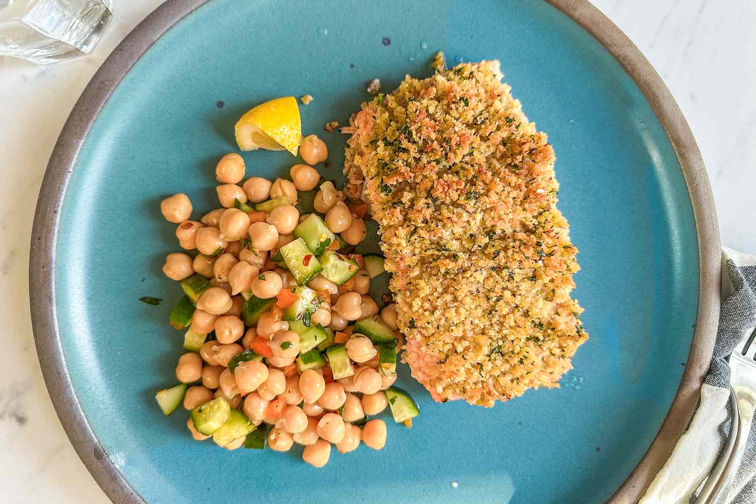 Overhead shot of a breaded salmon fillet next to chickpea salad on a blue plate