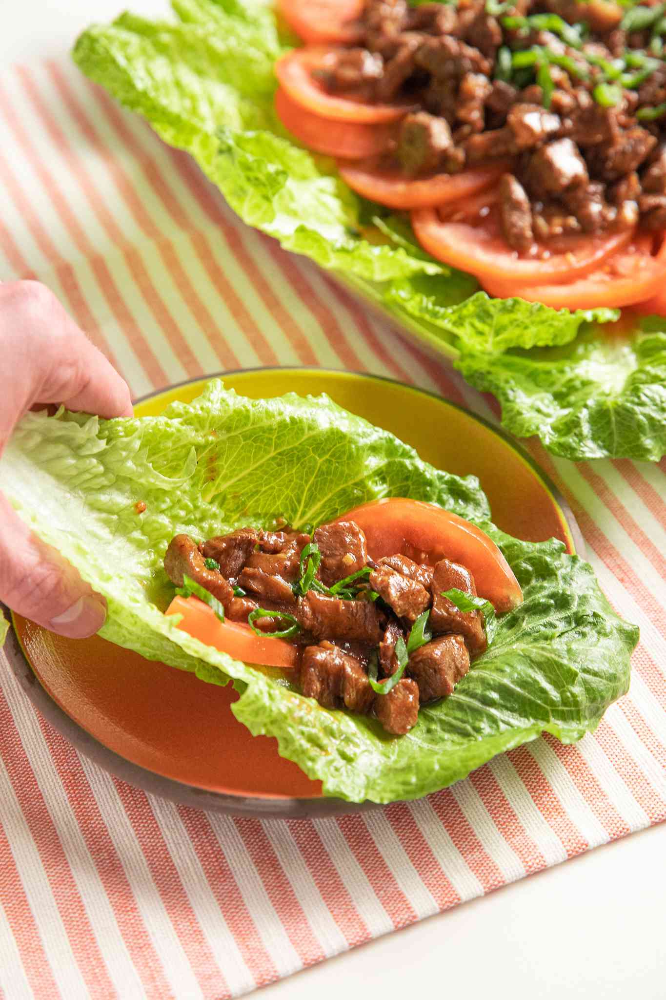 Hand Raising a Lettuce Leaf With a Bit of Lok Lak Over and a Sliced Tomato, and in the Surrounding, a Bowl of Pepper-lime Sauce and a Platter With More Lok Lak