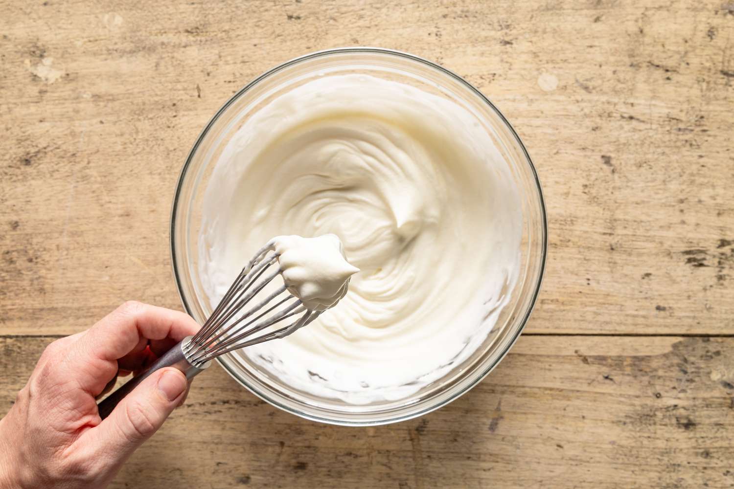 Overhead view of a hand holding a whisk over a clear glass bowl after whipping the cream topping for Company's Coming Pie recipe