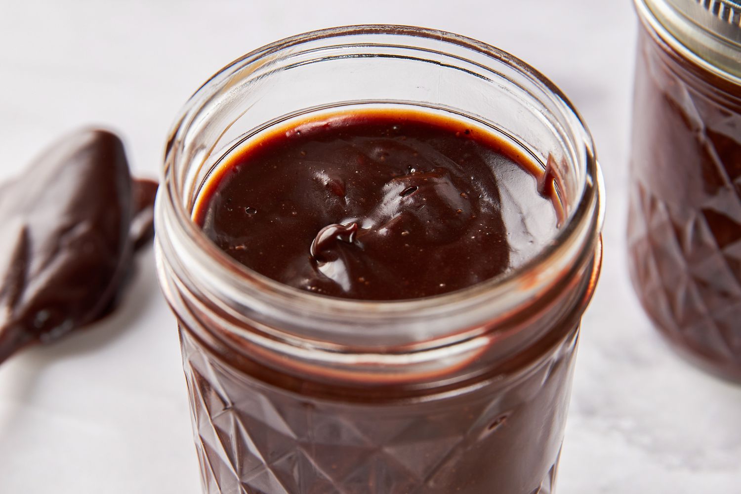 Hot Fudge Sauce in a pint mason jar next to another filled pint and a spoon