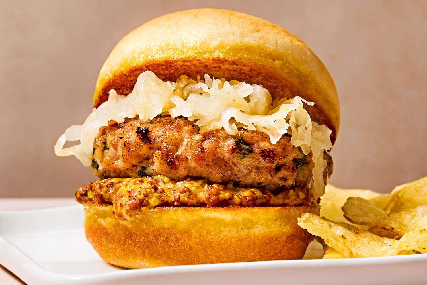 Closeup of a bratwurst slider topped with sauerkraut, on a plate with a side of potato chips