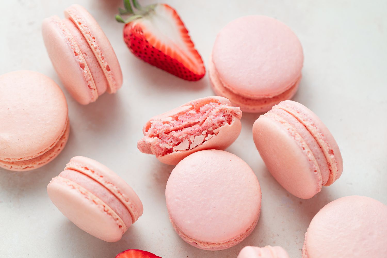 Overhead view of Strawberry macarons filled with strawberry buttercream scattered on a white background with the one in the center with a bite out of it.