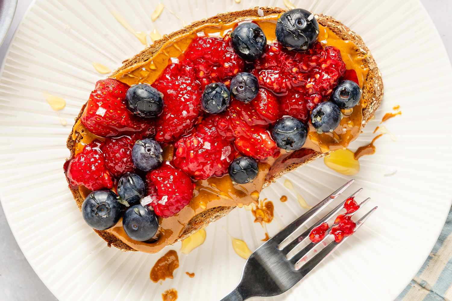 Slice of toast topped with peanut butter, raspberries, and blueberries, placed on a white plate with a fork to the side