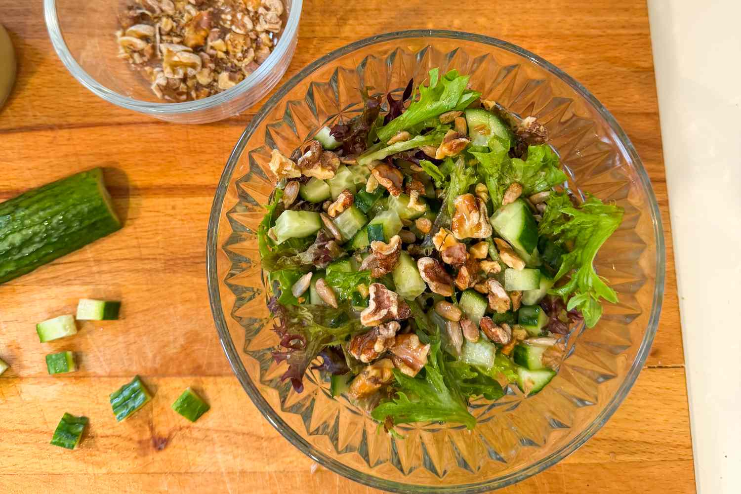A bowl of salad with greens and walnuts on a wooden surface accompanied by chopped cucumbers and scallions