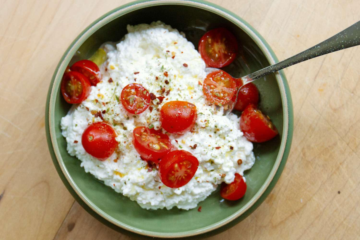 A green bowl containing cottage cheese topped with halved cherry tomatoes and seasoning with a spoon on the side