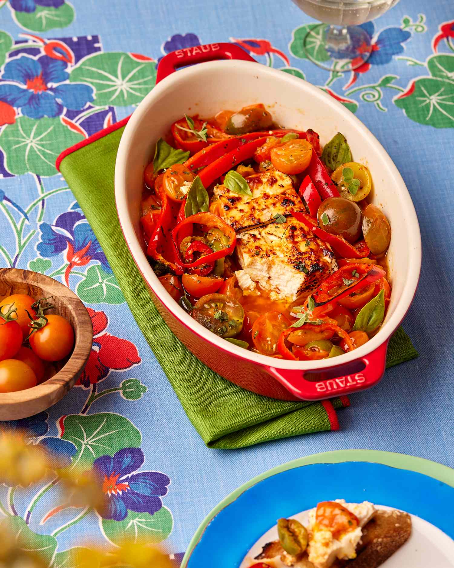 A red oval enameled cast iron baking dish with a block of feta in the middle surrounded by roasted peppers and tomatoes; the dish sits on a folded green napkin atop a colorful table cloth
